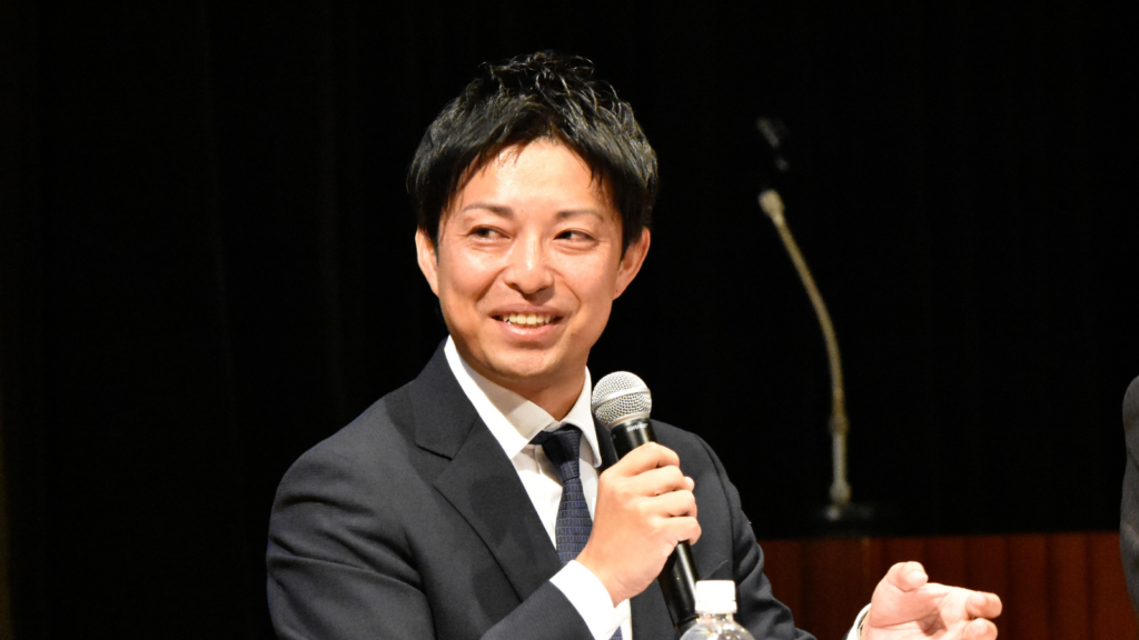 フォーラムでマイクを持って登壇している久志本組 6代⽬　代表取締役社⻑　清⽔良保 ⽒